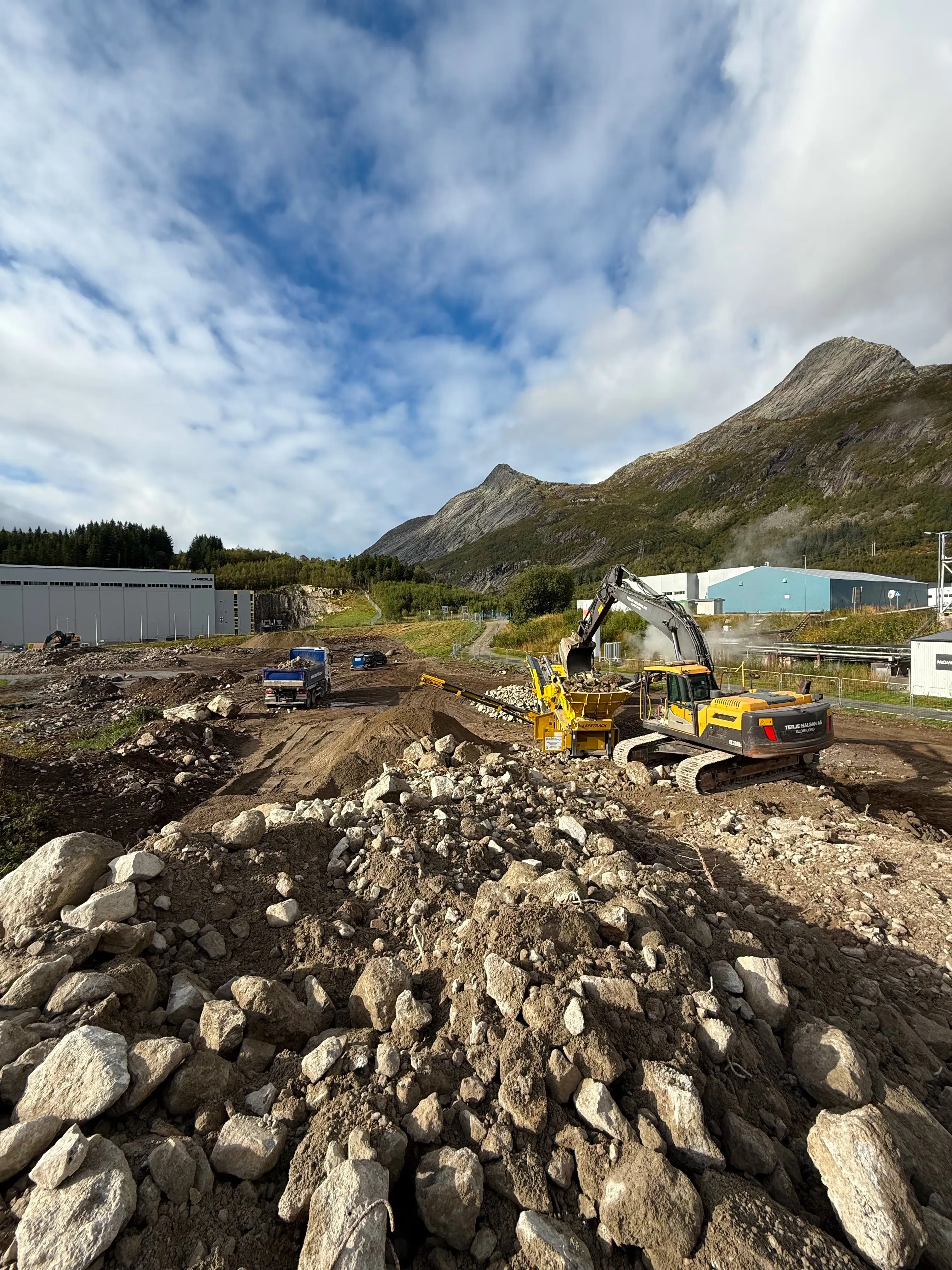 Gravemaskin og lastebiler på et anlegg med steinmasser, med fjell i bakgrunnen