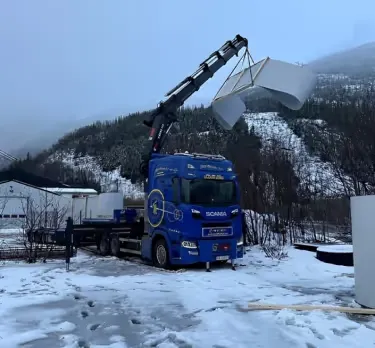 Blå lastebil med kran som løfter et element på en snødekt tomt