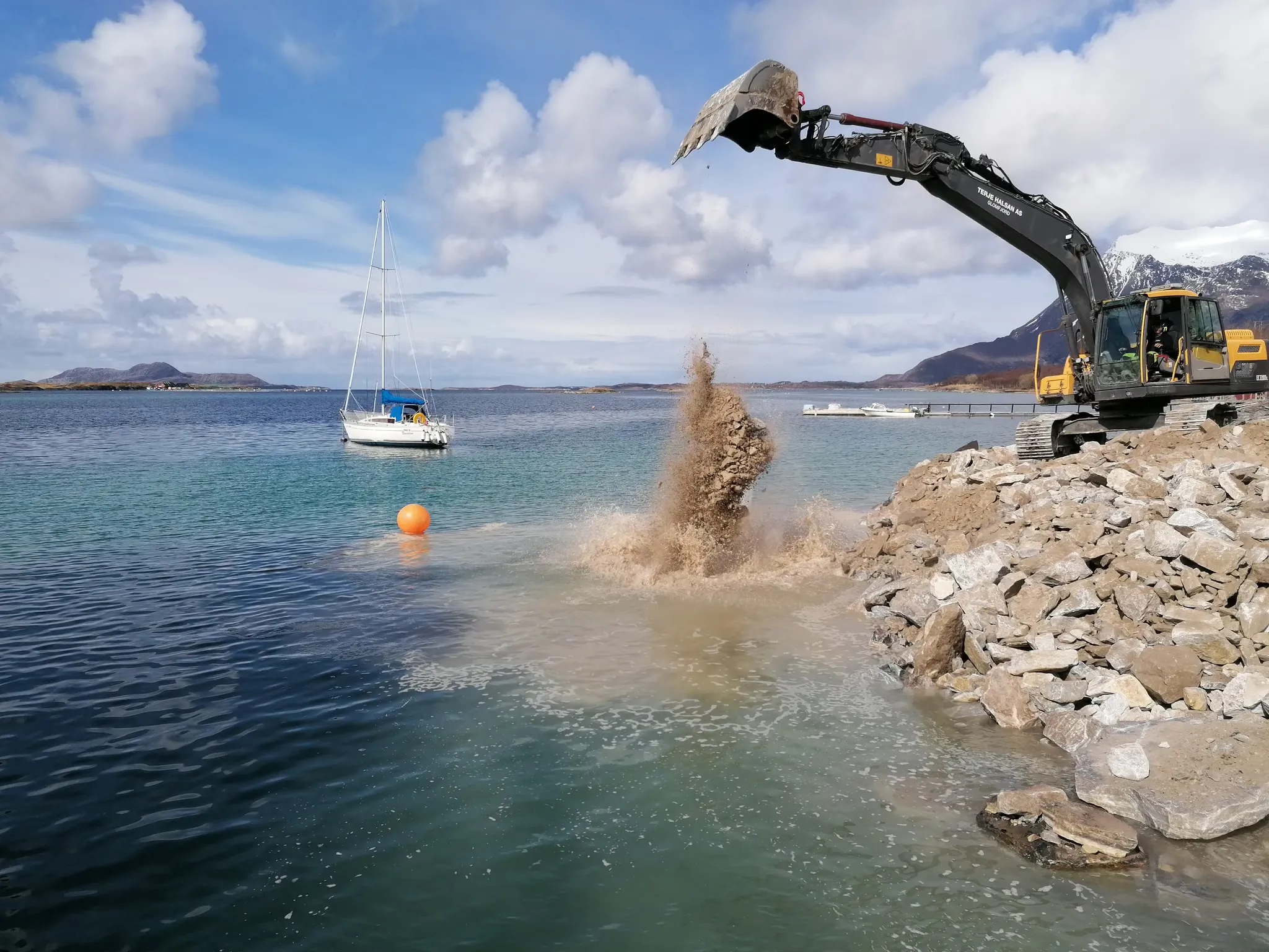 Gravemaskin som fyller stein og masse i sjøen, med en seilbåt bak