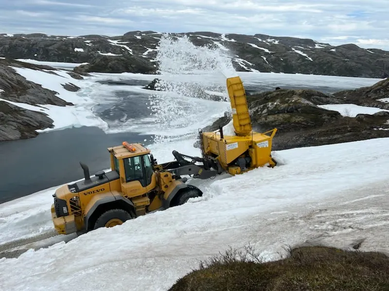 Gul Volvo-hjullaster med snøfreser som freser snø ved et islagt fjellvann