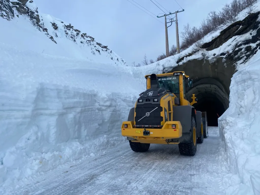 Gul hjullaster på en brøytet vei mellom høye snøvegger foran en tunnel