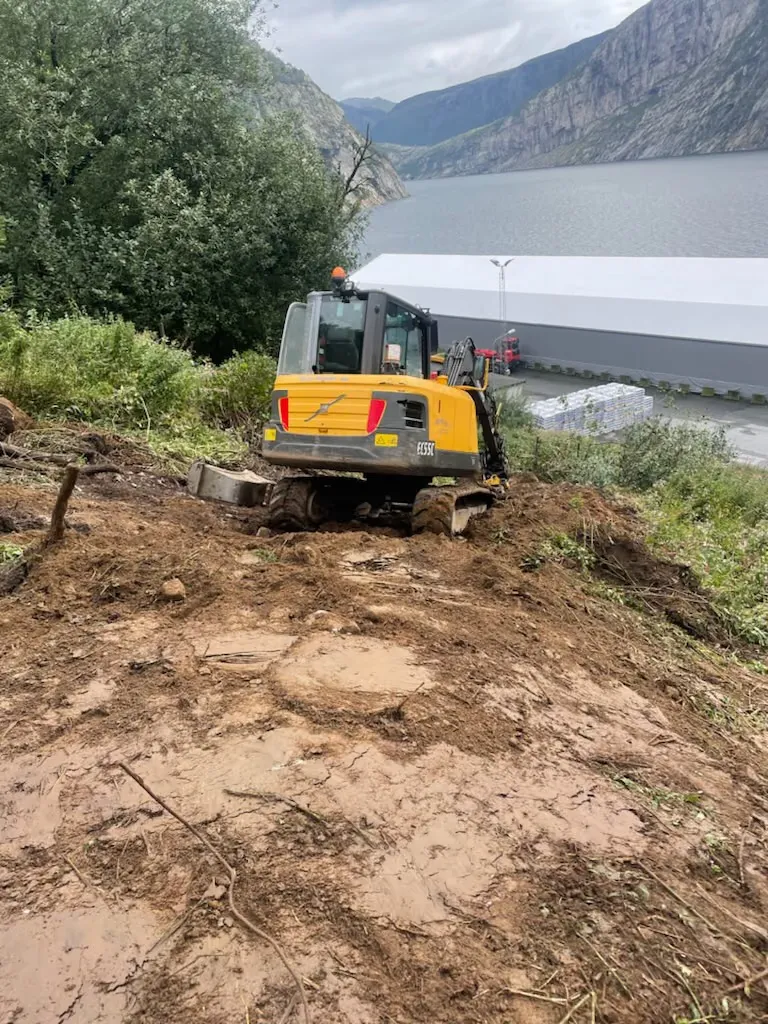 Gul gravemaskin som rydder en bratt skråning over fjorden