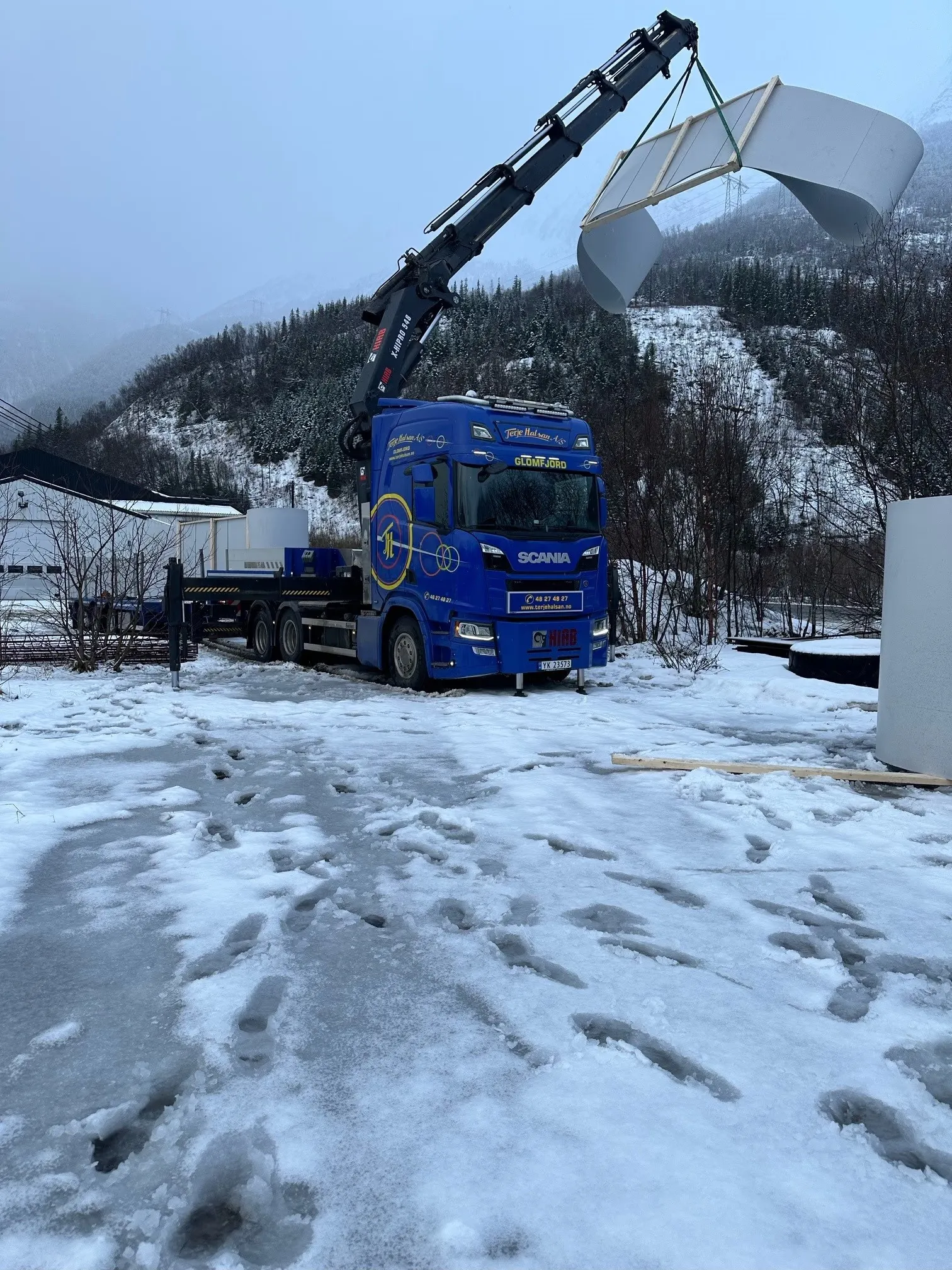 Blå lastebil med kran som løfter et element på en snødekt tomt