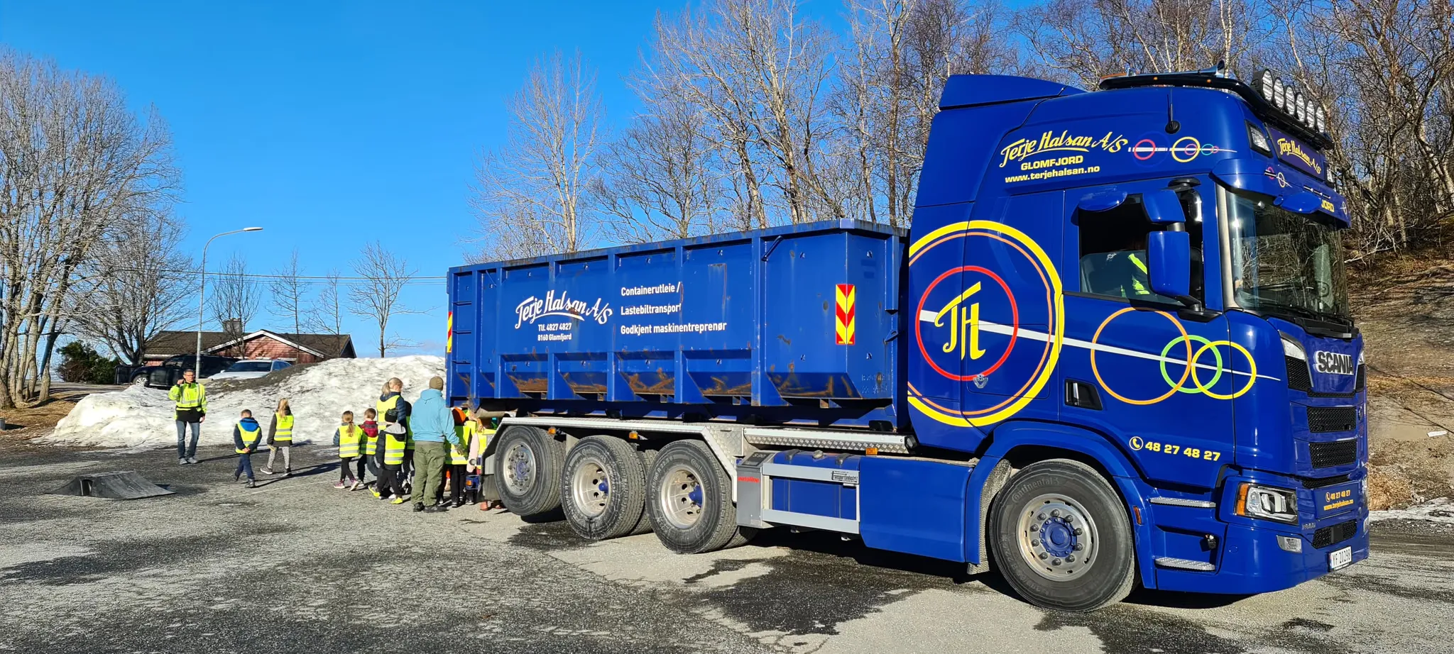 Blå Scania-krokbil med container, med en gruppe barn i refleksvester ved siden av