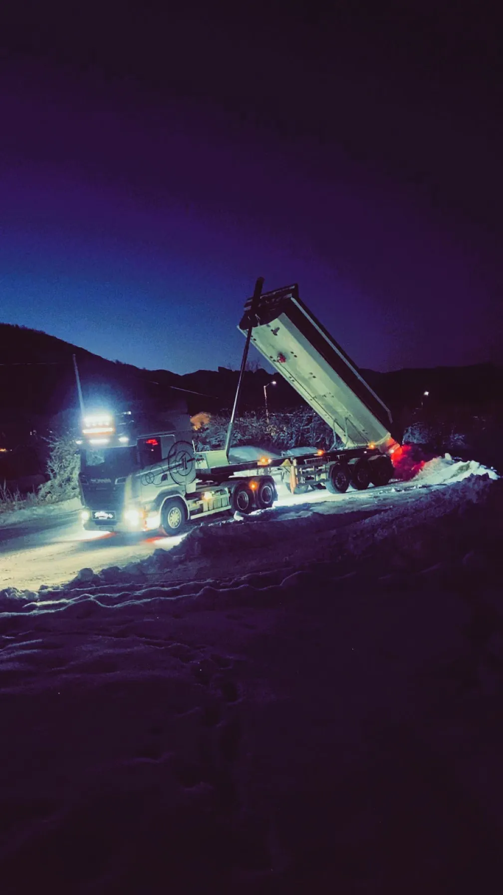 Tippsemitrailer som tipper i snøen om kvelden, opplyst av arbeidslys