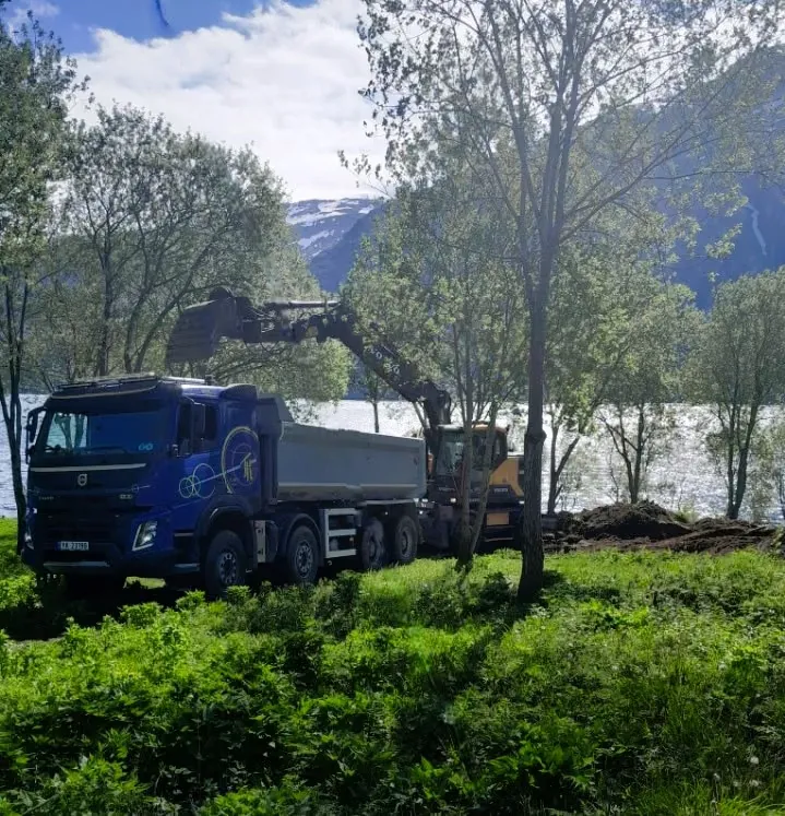 Blå Volvo-tippbil som lastes av en gravemaskin mellom trær ved sjøen