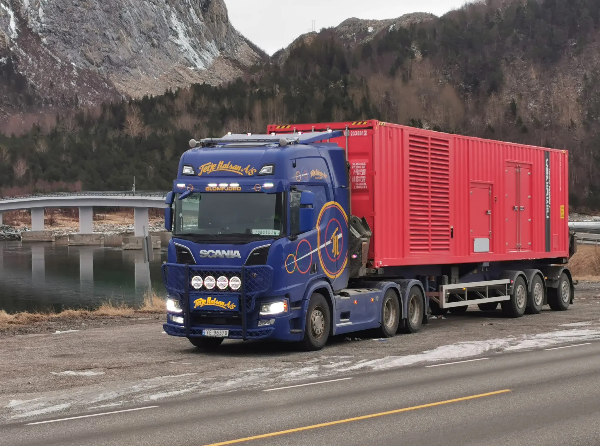 Blå Scania-trekkvogn med rød container på semitrailer, med bro og fjell bak