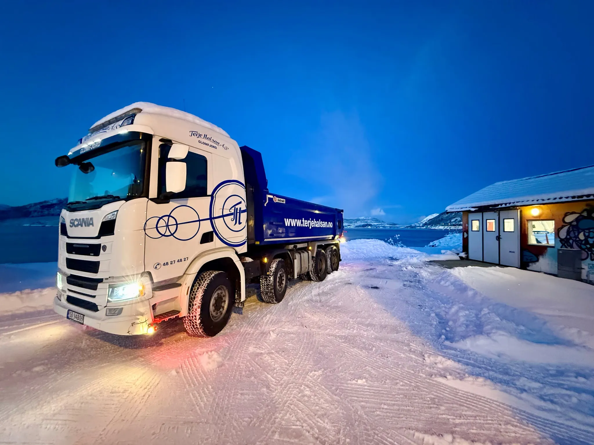 Hvit Scania-tippbil med blått lasteplan på en snødekt plass i blåtimen