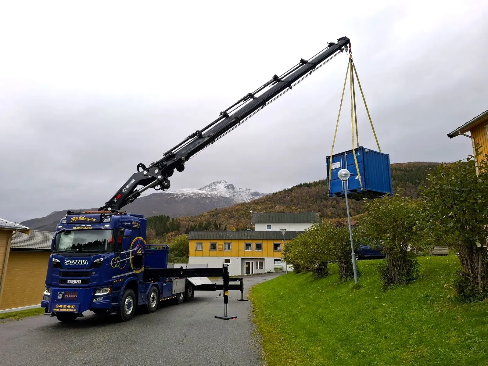Blå Scania-lastebil med kran som løfter en blå container ved gule bygninger