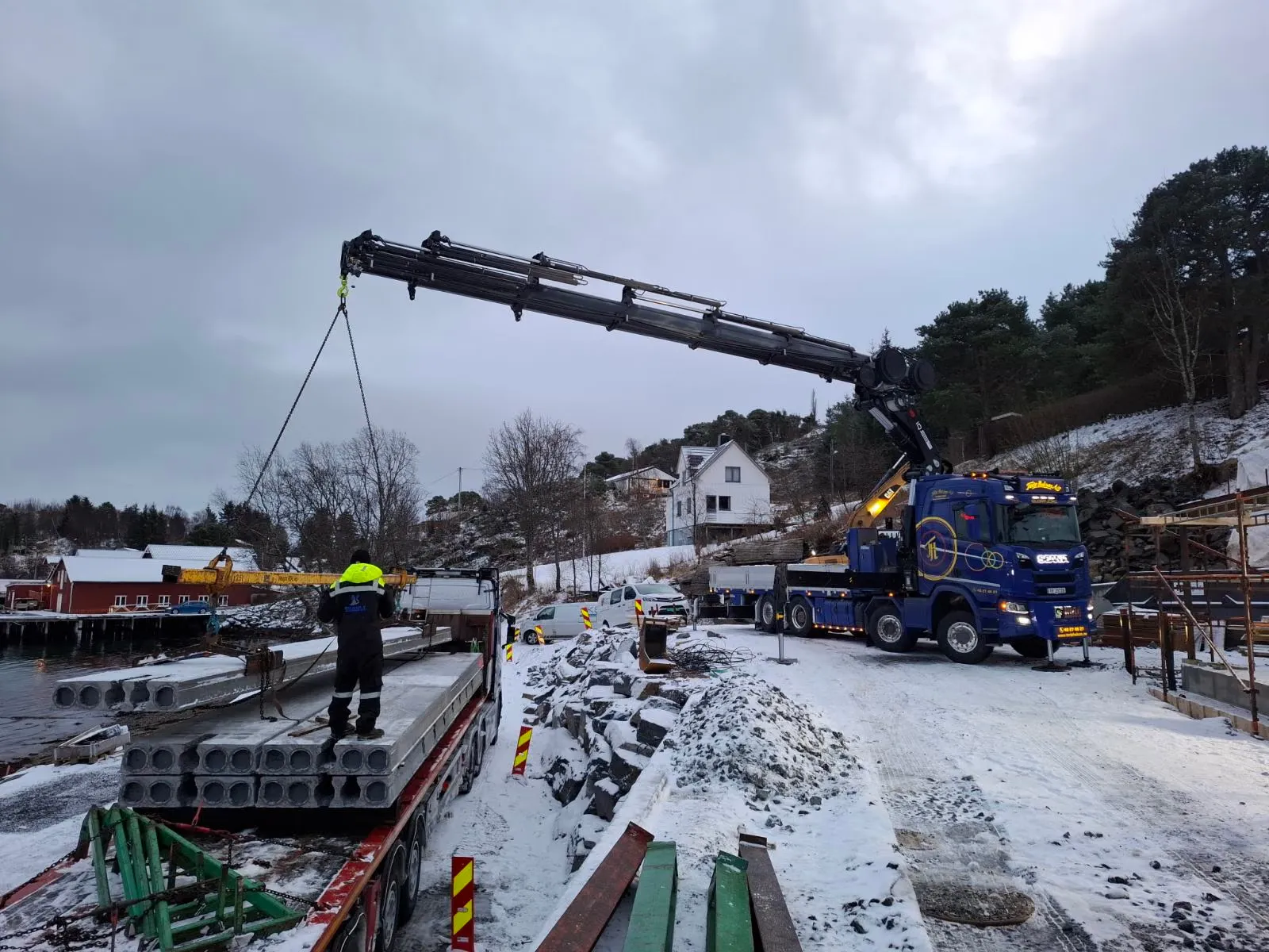 Kranbil som løfter betongelementer av en tilhenger ved sjøen om vinteren