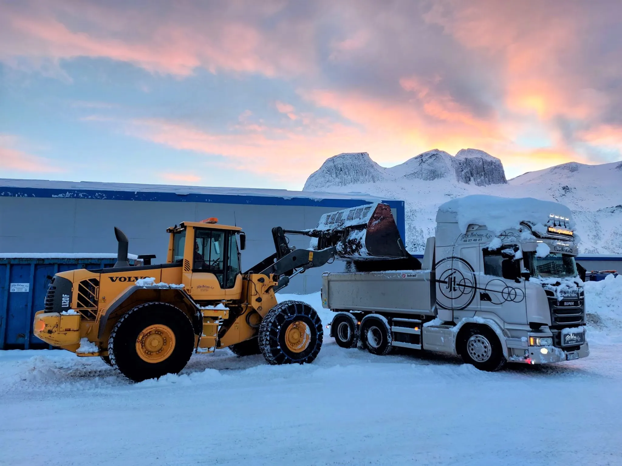 Gul Volvo-hjullaster som laster en hvit Scania-tippbil foran snødekte fjell i solnedgang