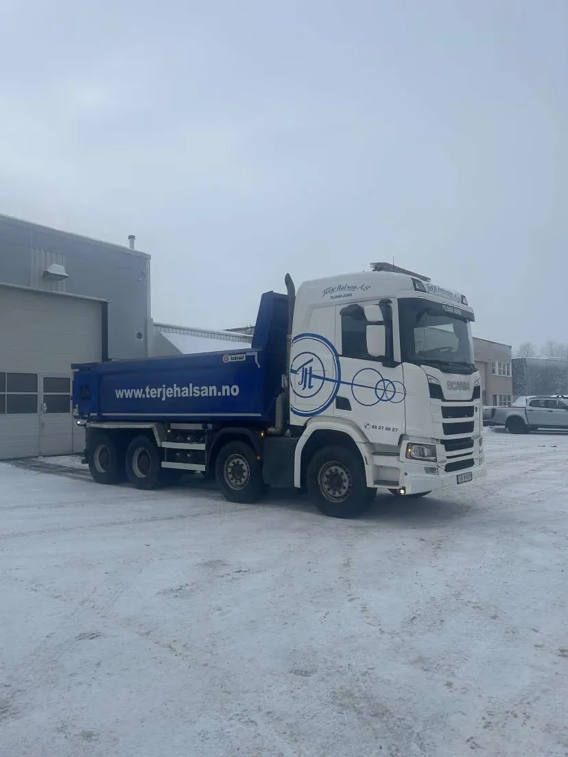 Hvit Scania-tippbil med blått lasteplan utenfor en lagerhall i snøen