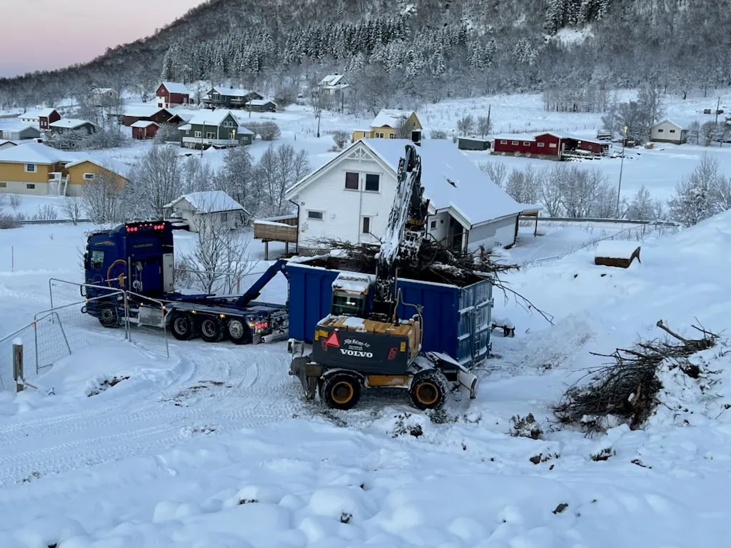 Blå krokbil og gul Volvo-gravemaskin som laster kvist i en container i et snødekt boligfelt